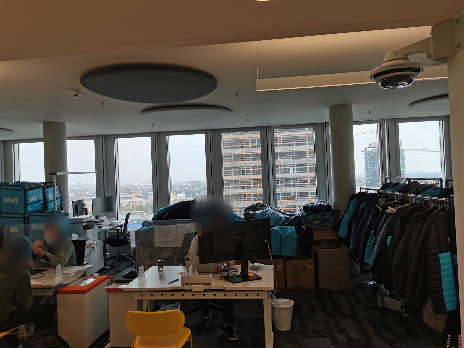 The Wolt Berlin office in daily operation: desks in use, stacked thermal boxes, packed bags and jackets on rails along the window wall.