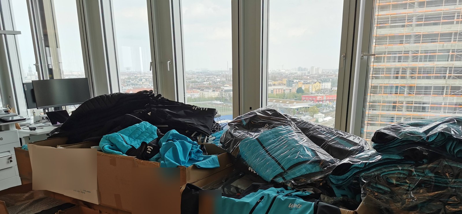 Cardboard boxes and stacked courier jackets and thermal bags piled against the windows of the Wolt Berlin office, without shelving or intake system.