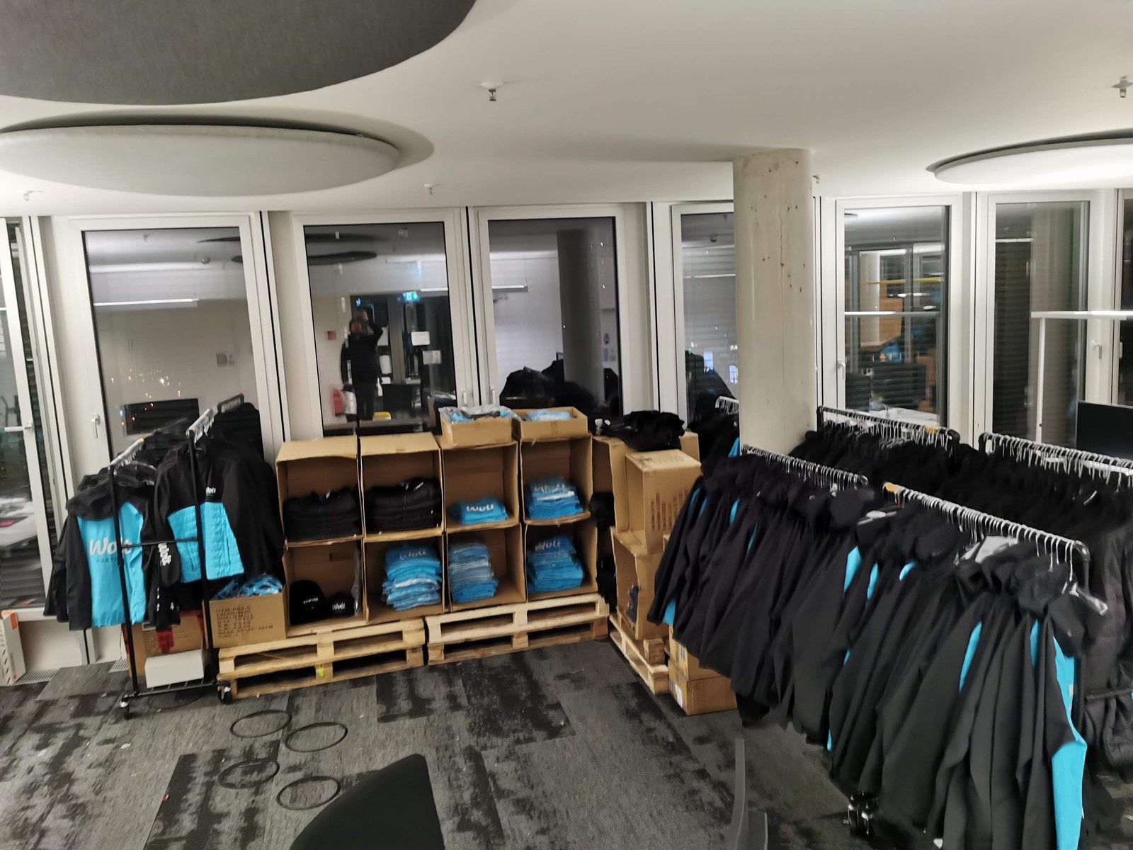 Distribution station at the Wolt Berlin office: jackets on rolling rails, folded shirts sorted by size in open cases, and pallets of boxed stock.
